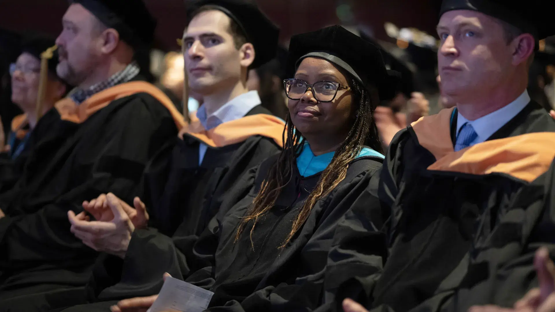 A woman wearing graduation robes sits in a crowd of people 
