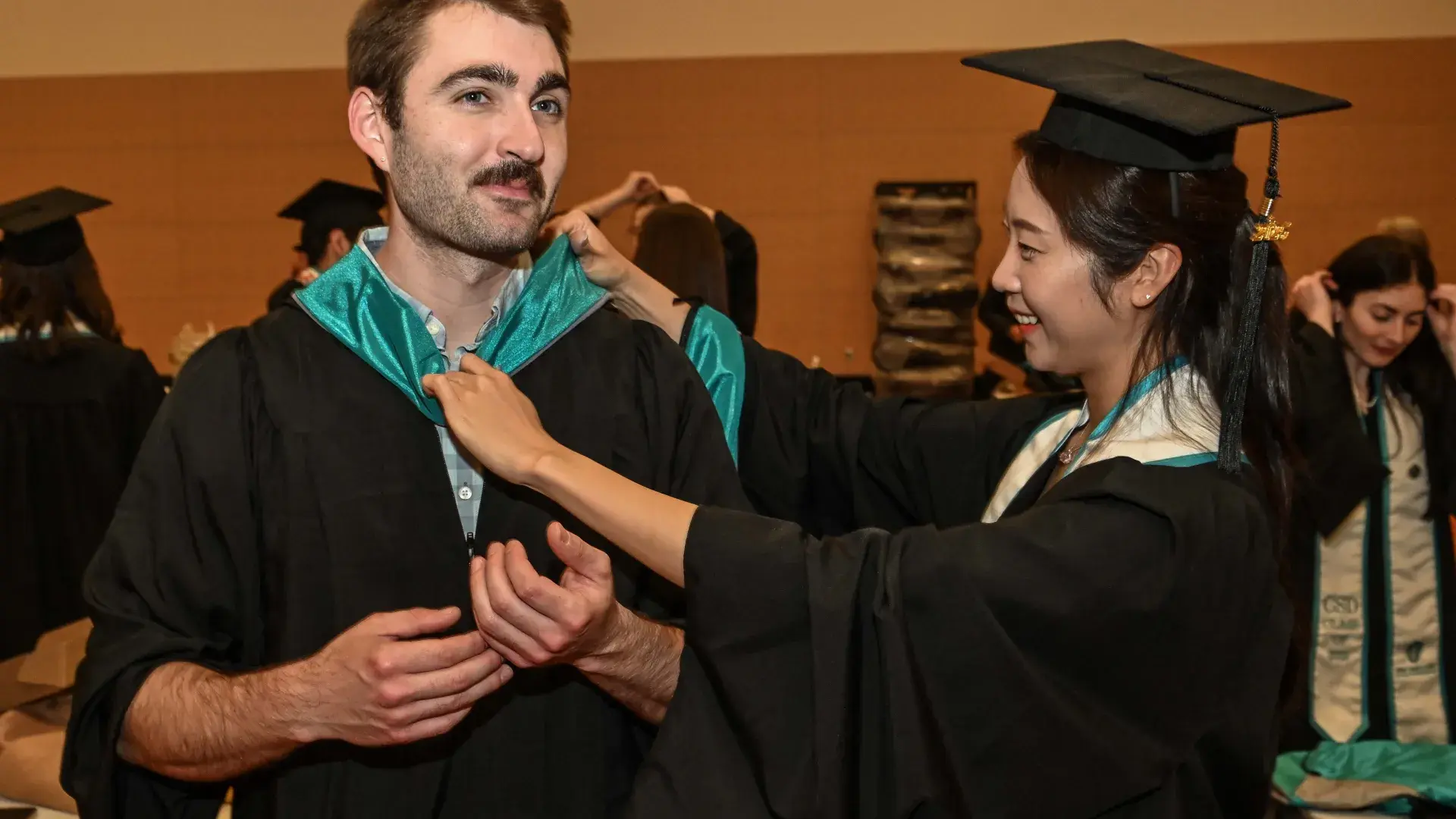 A man gets his blue hood adjusted on his graduation gown from a woman with a cap and gown on as well. 
