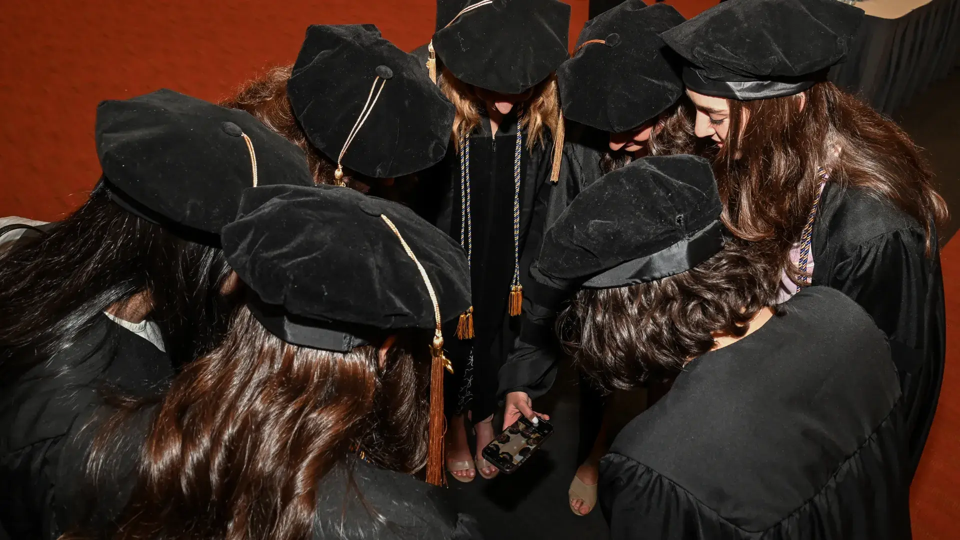 7 students are looking down at a phone in a circle, taking a photo. 