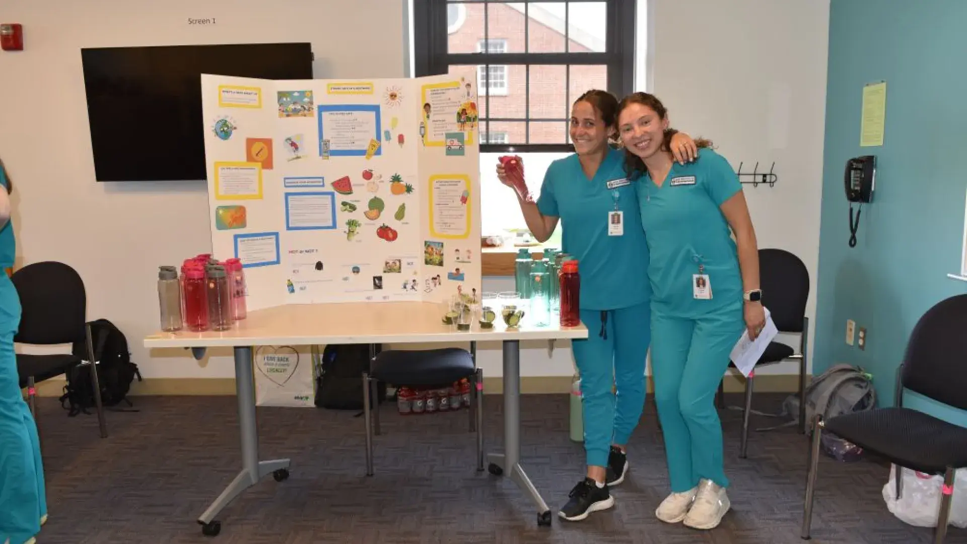 Two women wearing medical scrubs pose in front of a display board and table