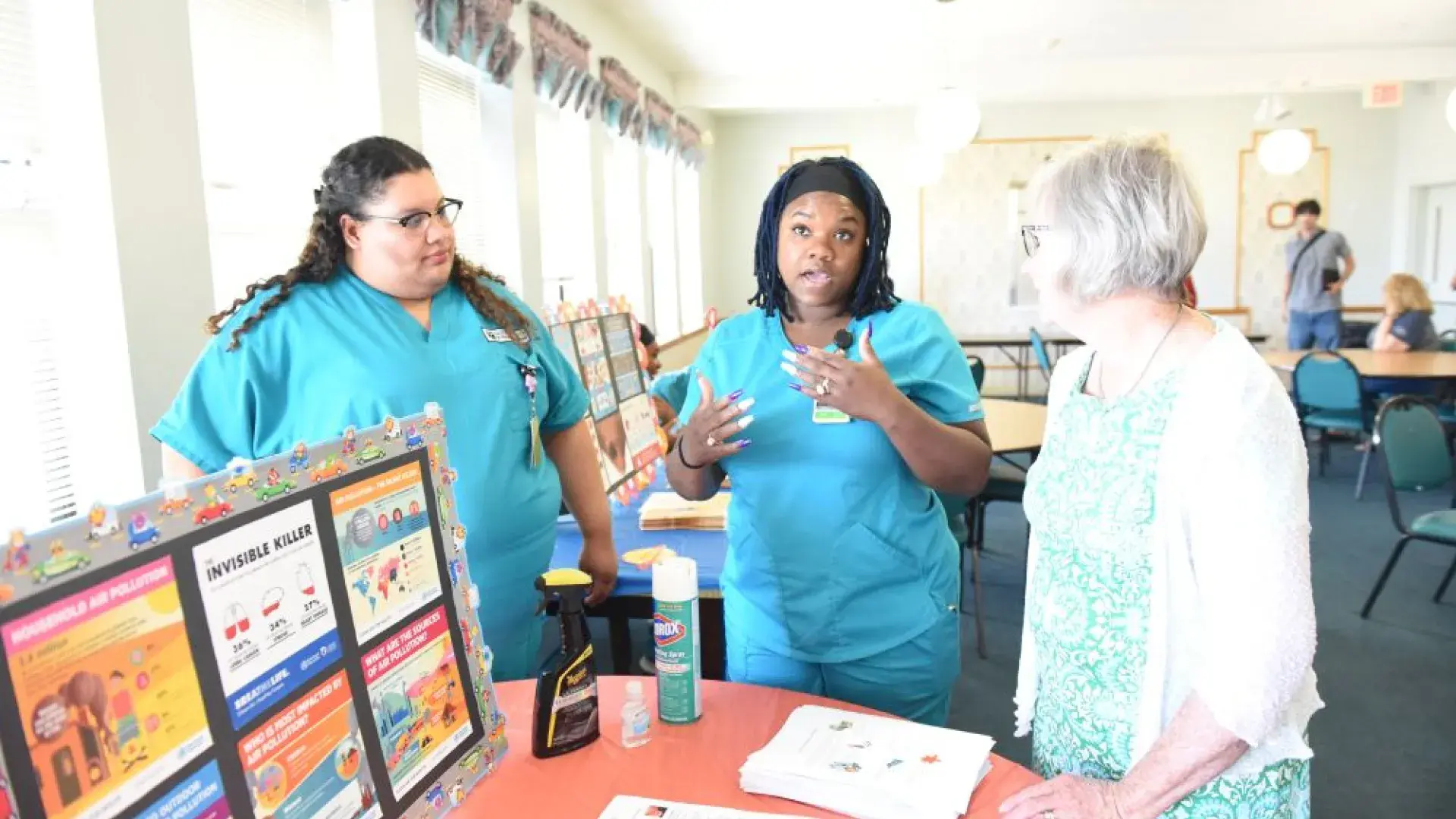 Two women wearing medical scrubs talk to a third woman near a table with a display