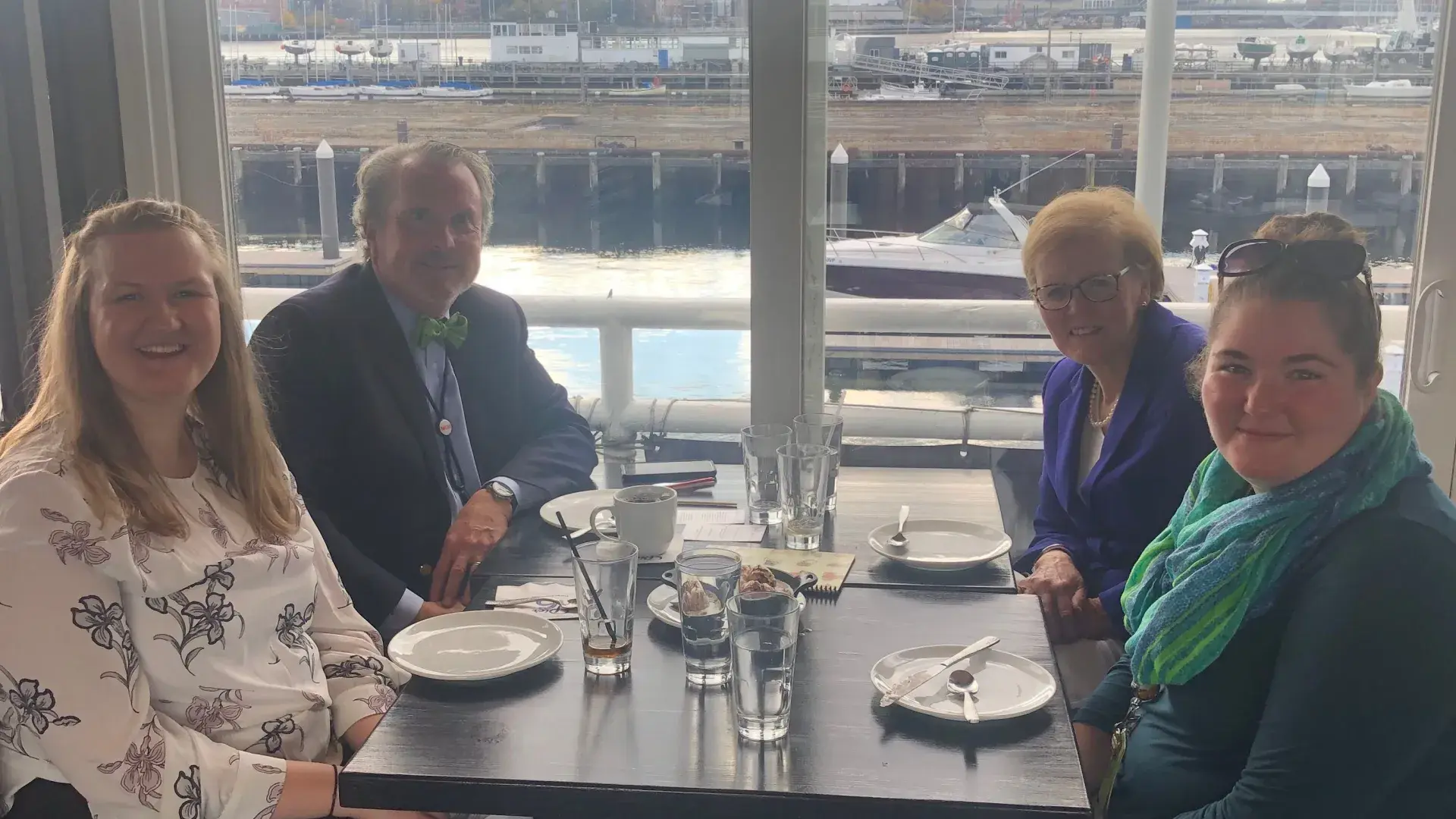 Four people sit at a restaurant table with a view of the sea through the window behind them