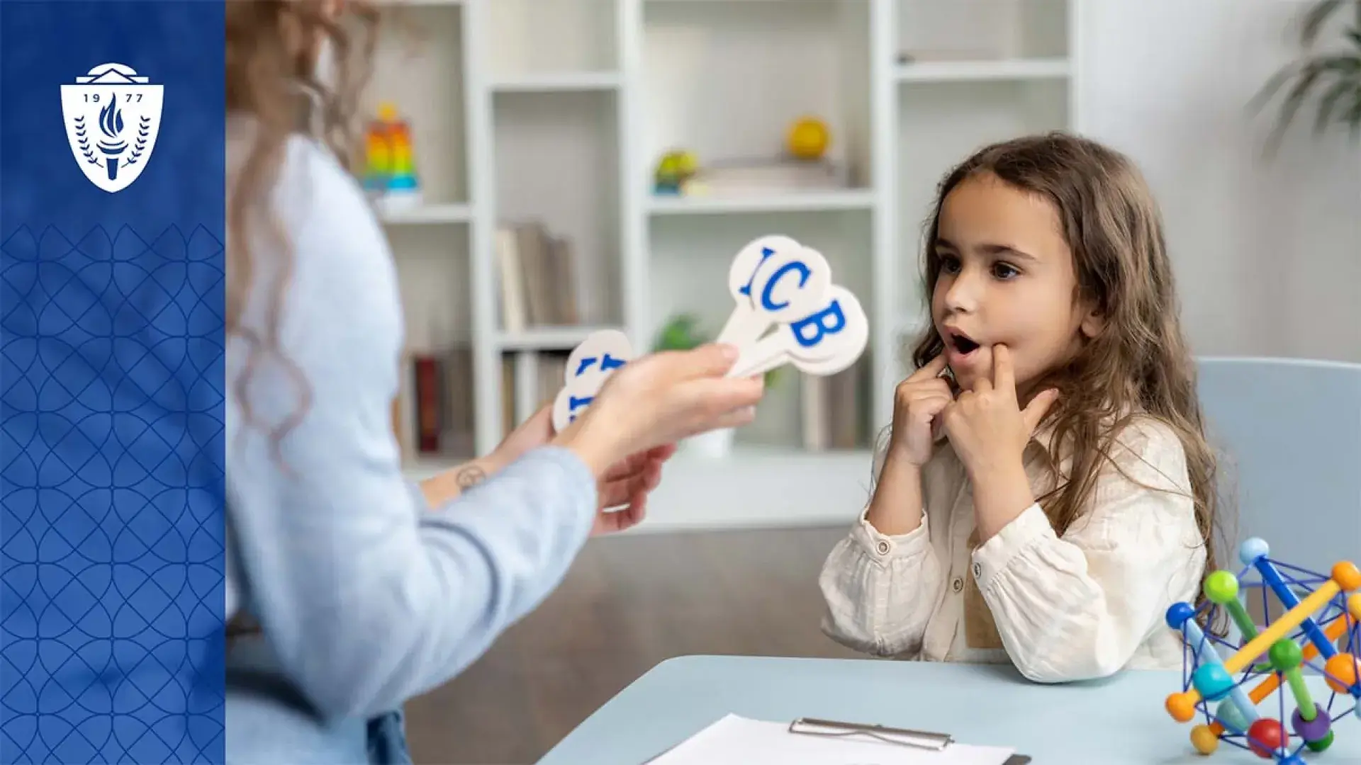 Young girl sitting at desk looking at a teacher holding sticks with alphabet letters on them