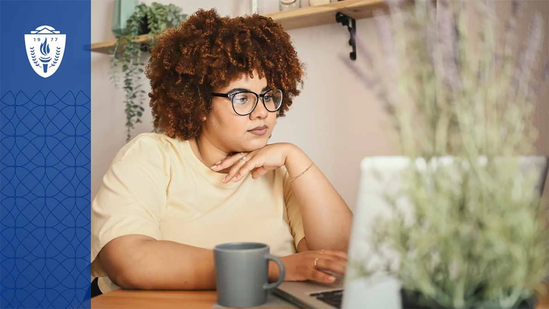 Woman wearing a yellow shirt sitting at a desk working on a laptop next to a coffee mug