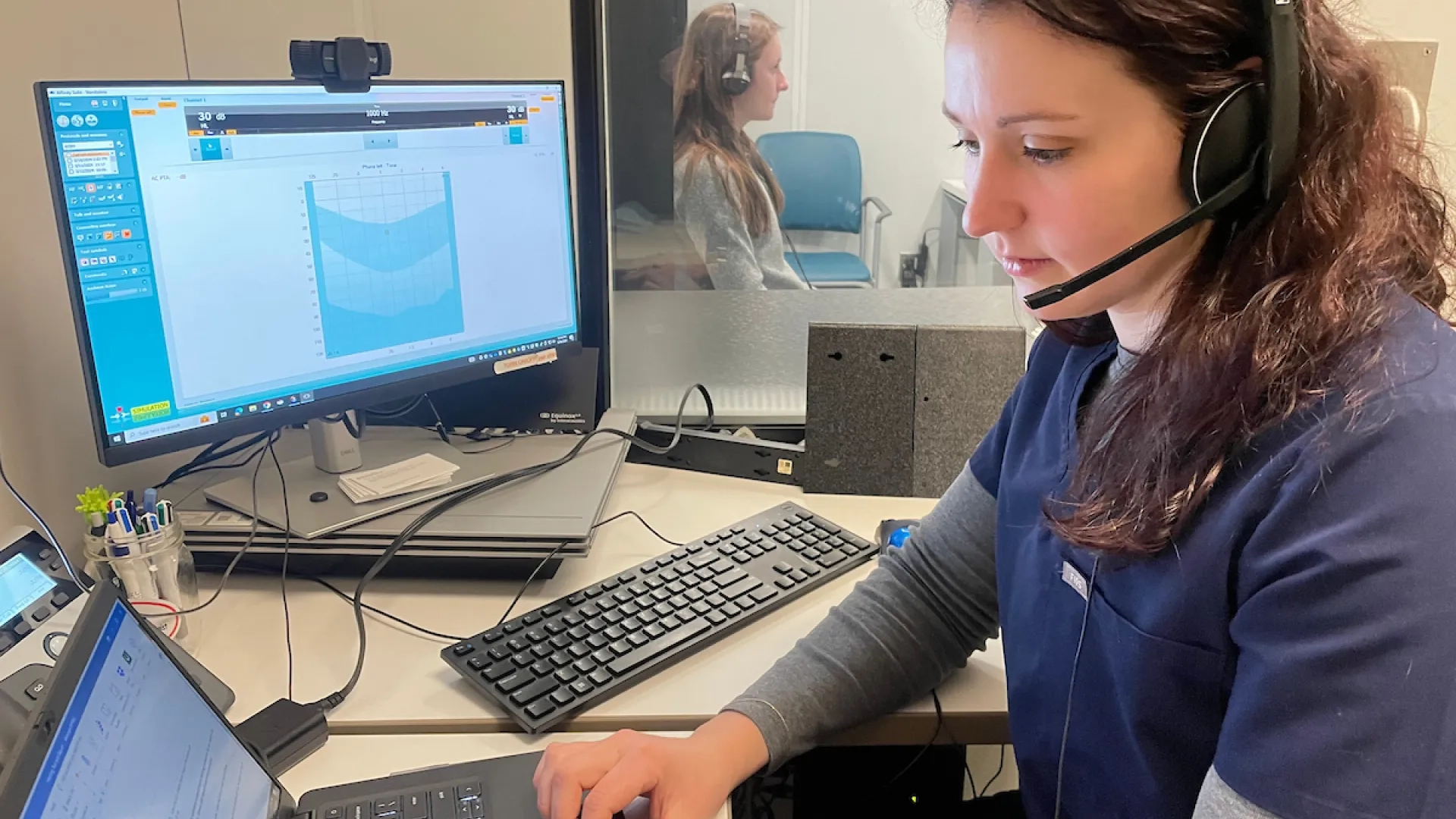 woman wearing headset looks at computer while a woman is viewable behind a glass window in the next room