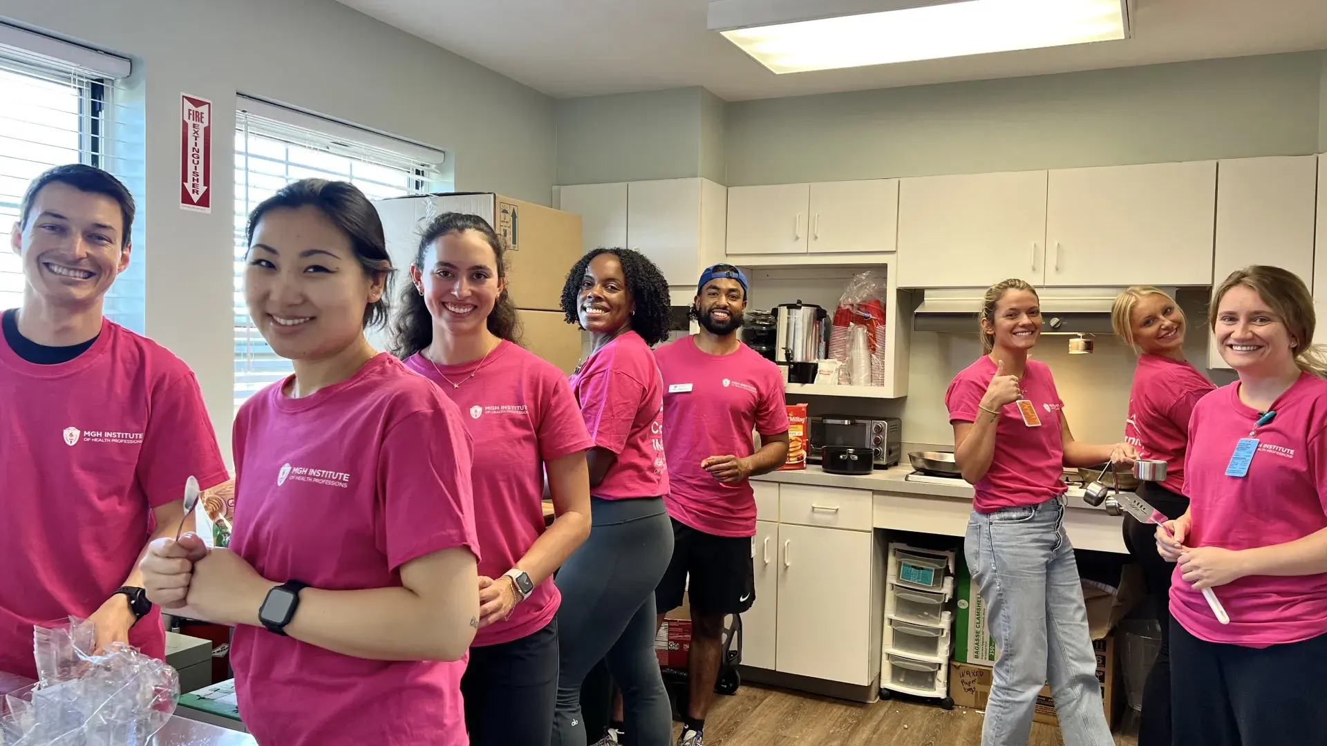Eight people stand in a kitchen wearing pink shirts