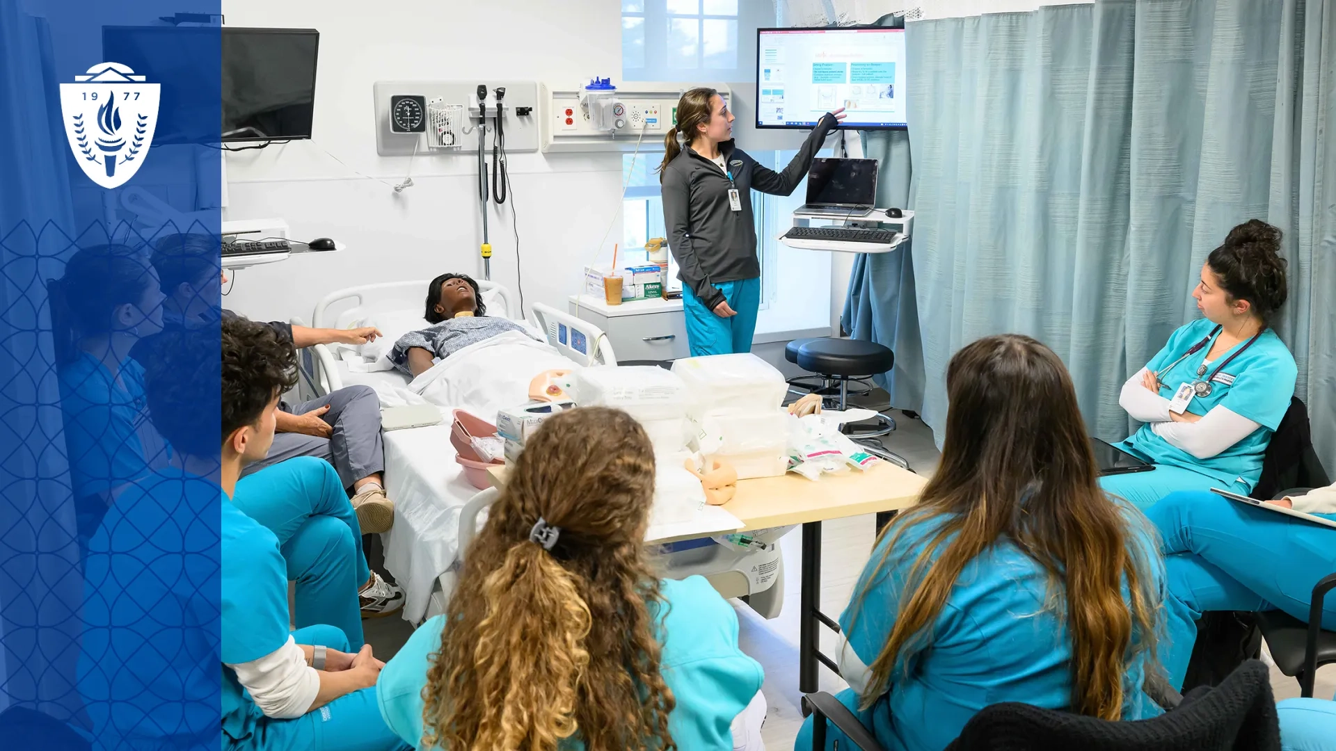 Group of students seated around simulation hospital bed with a mannequin laying on it. An instructor points toward the computer screen.