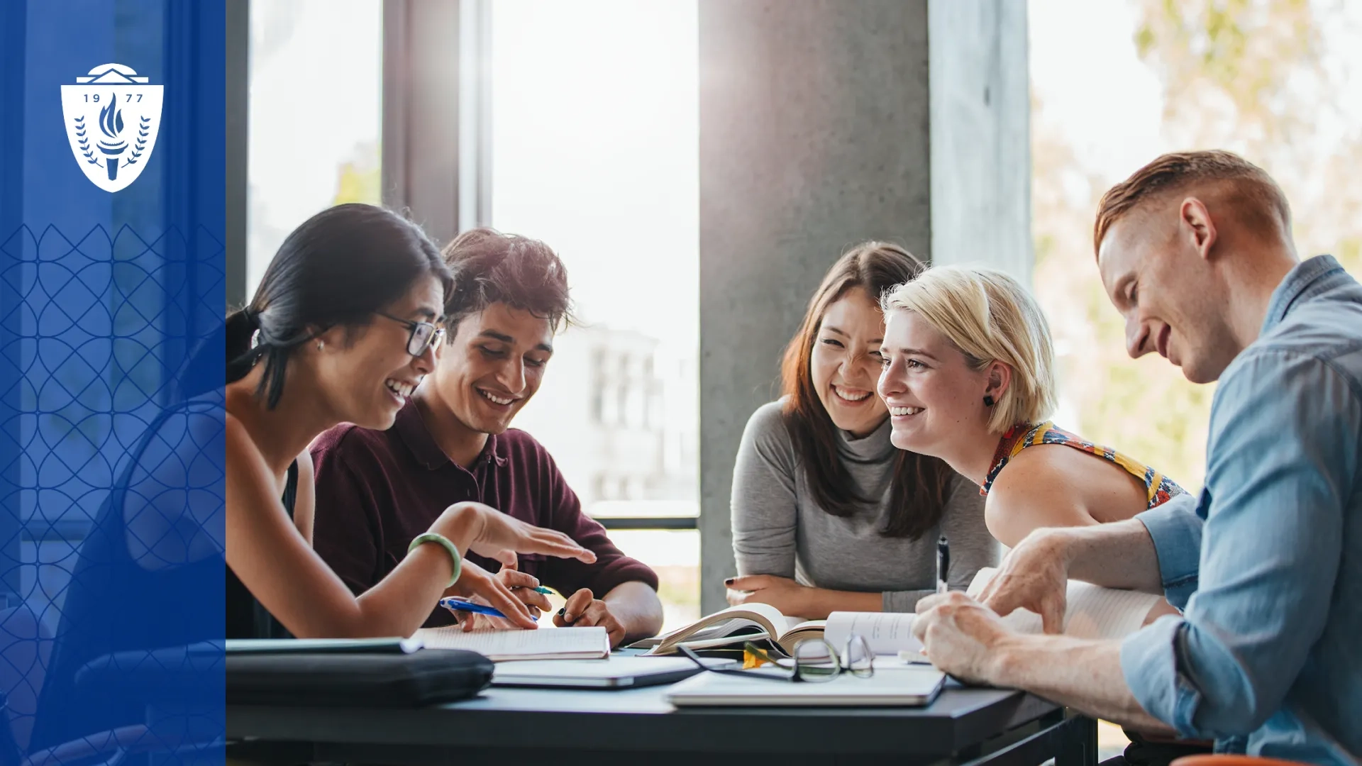 Group of students sitting around desk smiling, reading books, and taking notes.