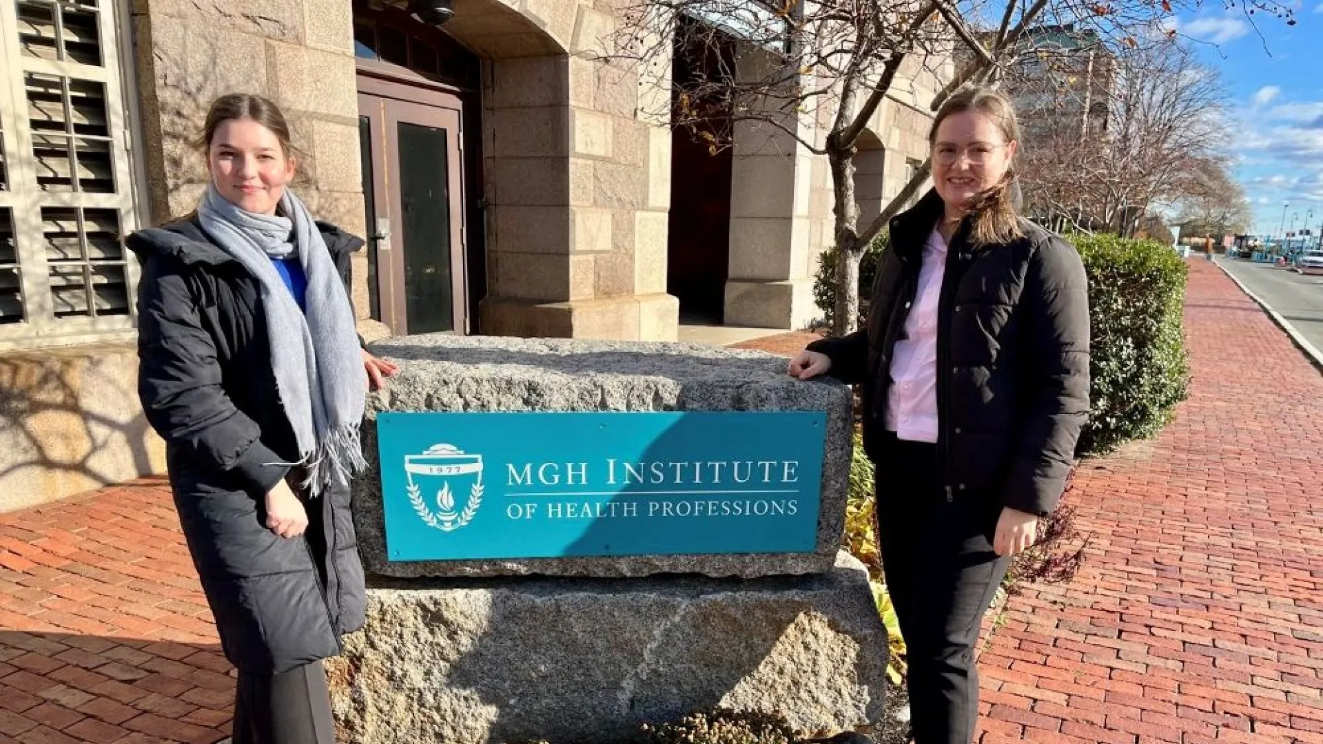 Two women pose on either side of the University's sign in front of the Shouse Building
