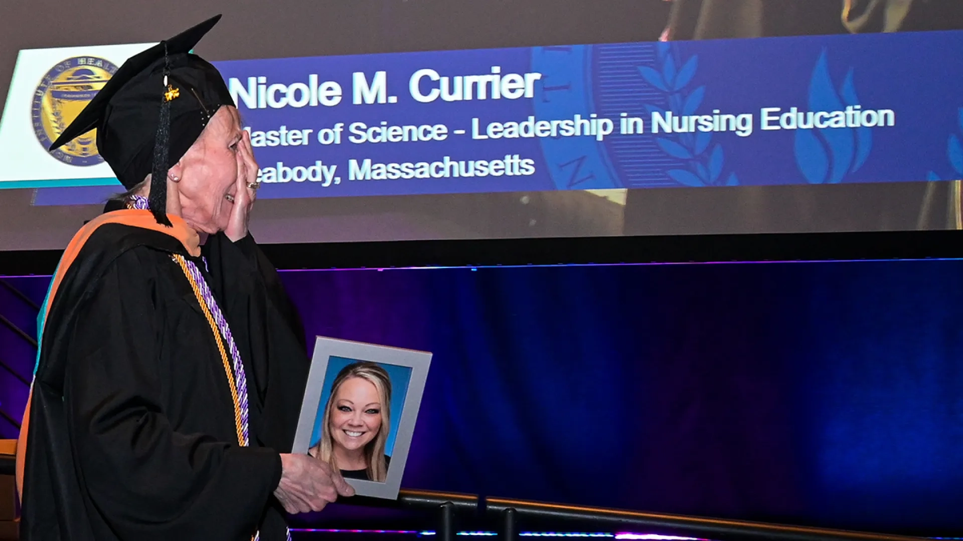 A woman walks across a commencement stage holding a photo of a young woman with a screen in the background with the name Nicole M. Currier