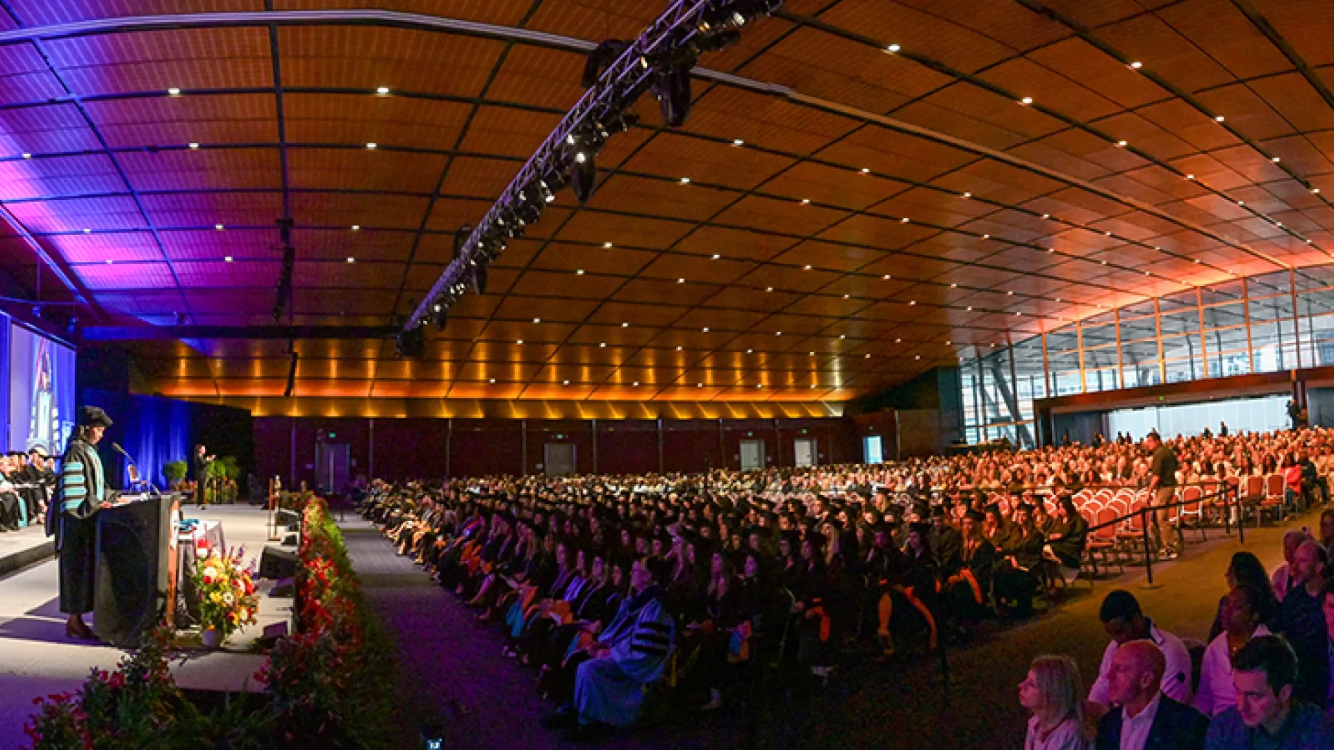 A large room with people dressed for commencement and an audience behind them