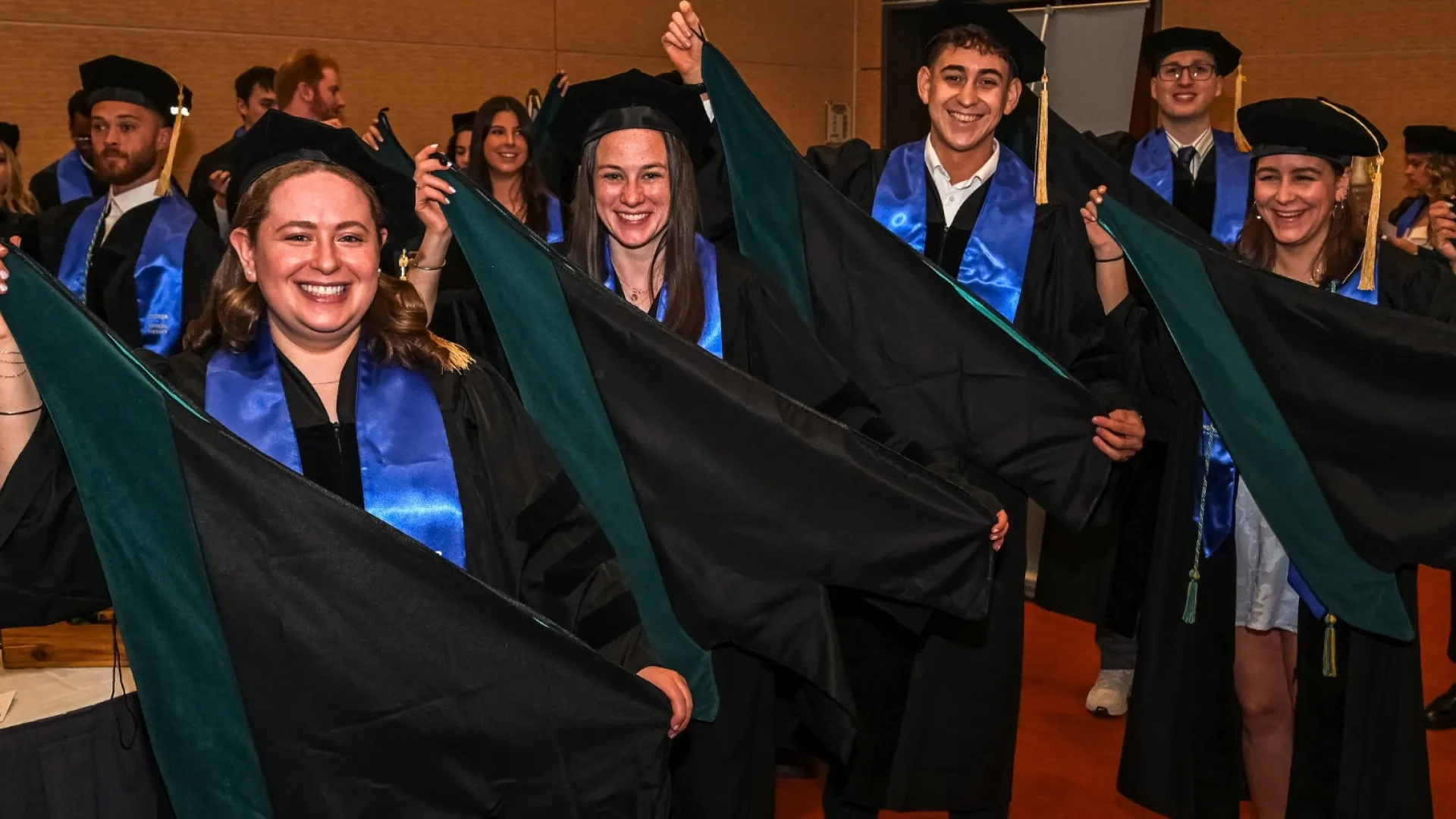 students in graduation attire hold up pieces of fabric that will become their "hoods"