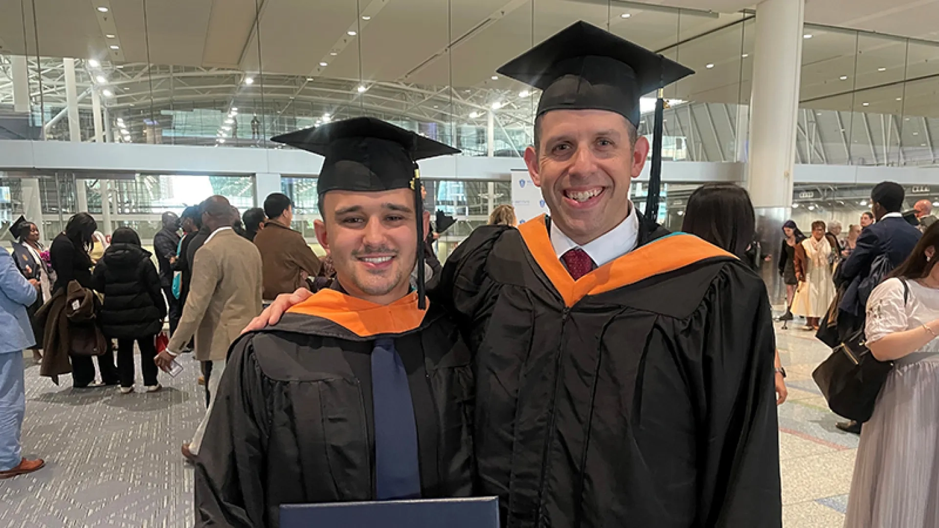 Two men wearing graduation gowns and caps. One is holding a diploma