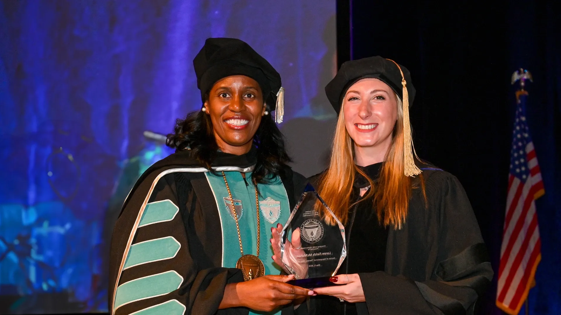 Two women in graduation clothing holding an alumni award