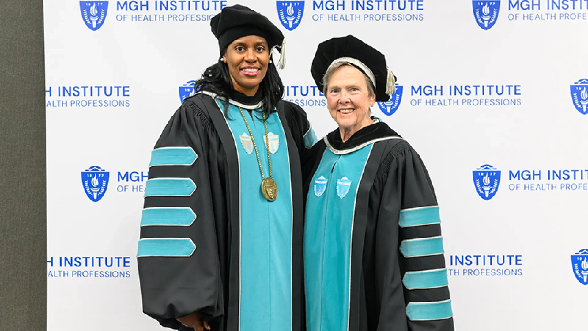 Two women in graduation gear pose in front of a backdrop that says MGH Institute of Health Professions