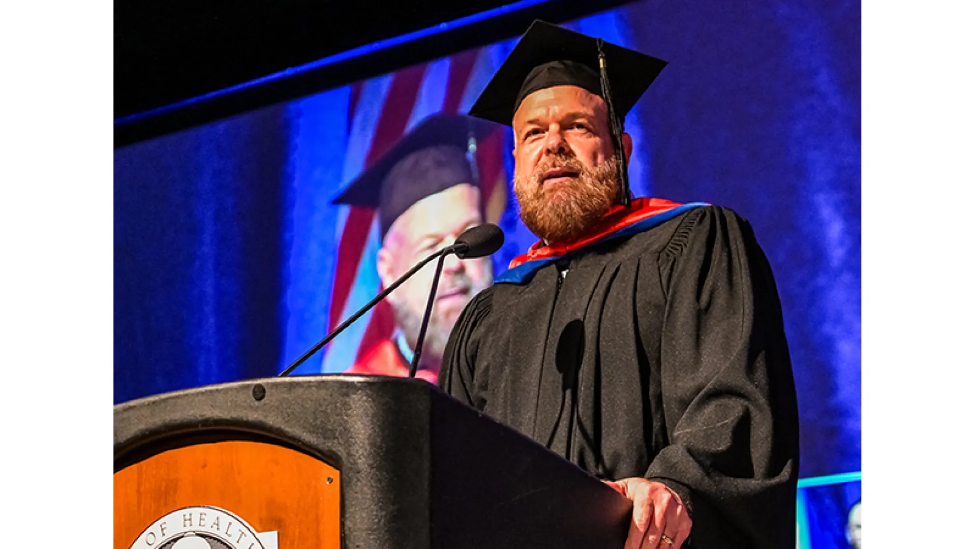 A man stands at a podium wearing graduation robes at the 2026 commencement  
