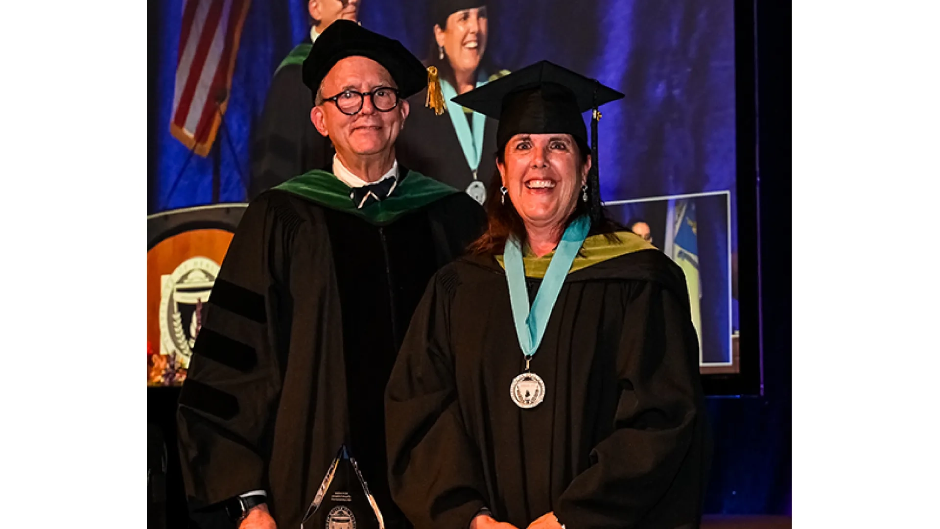 Two people in graduation robes, one wearing a medal, stand on a stage