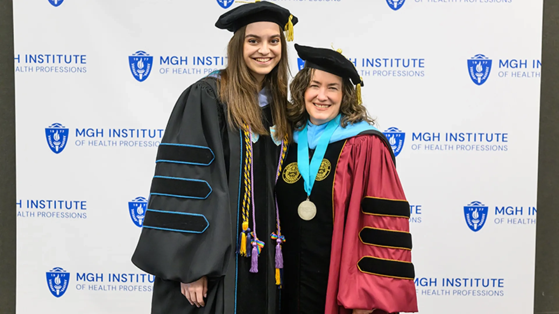 Two women in graduation gear pose in front of a backdrop that says MGH Institute of Health Professions