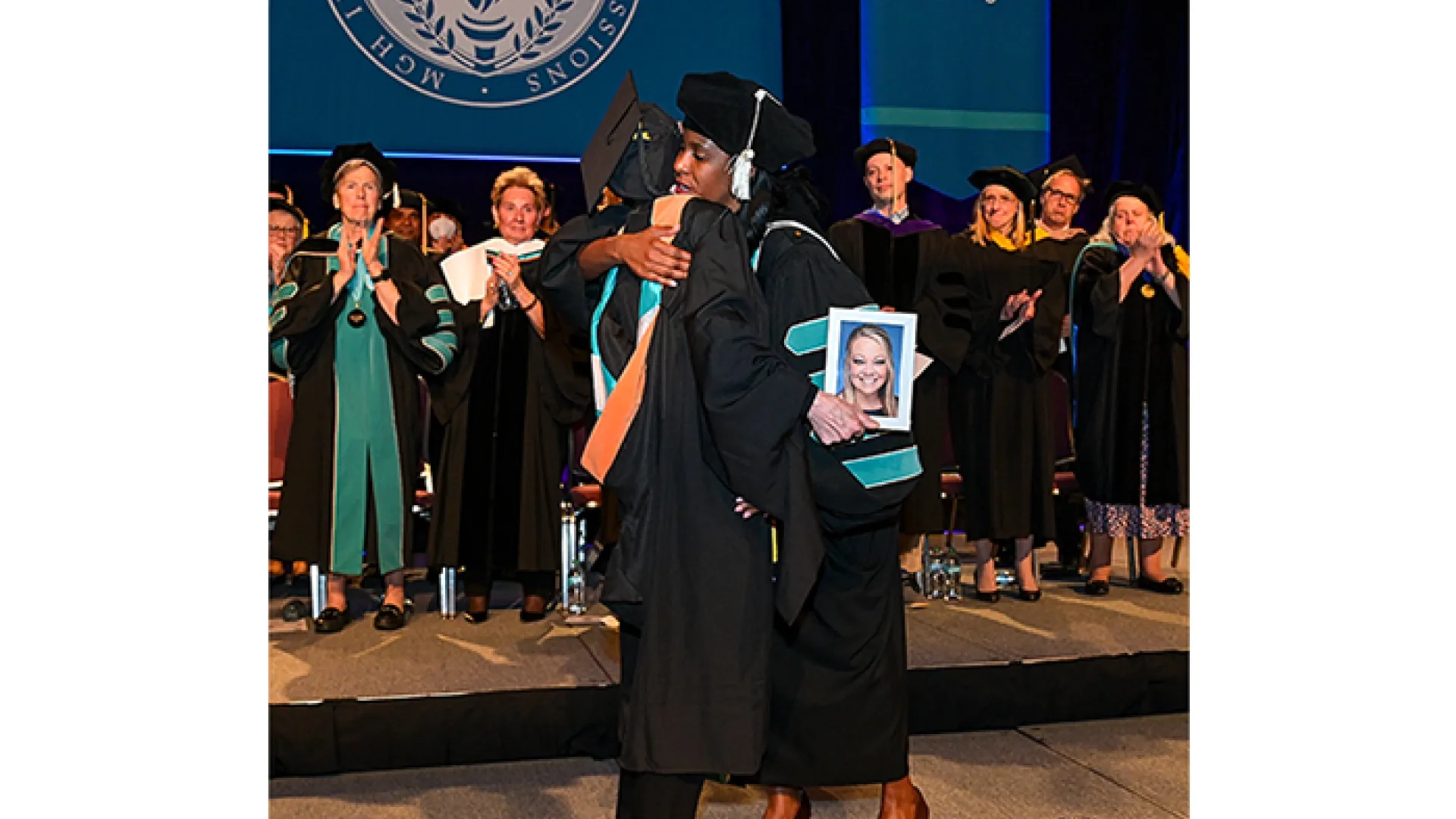 Two people in graduation gowns hug on a stage while one holds a photo of a young woman