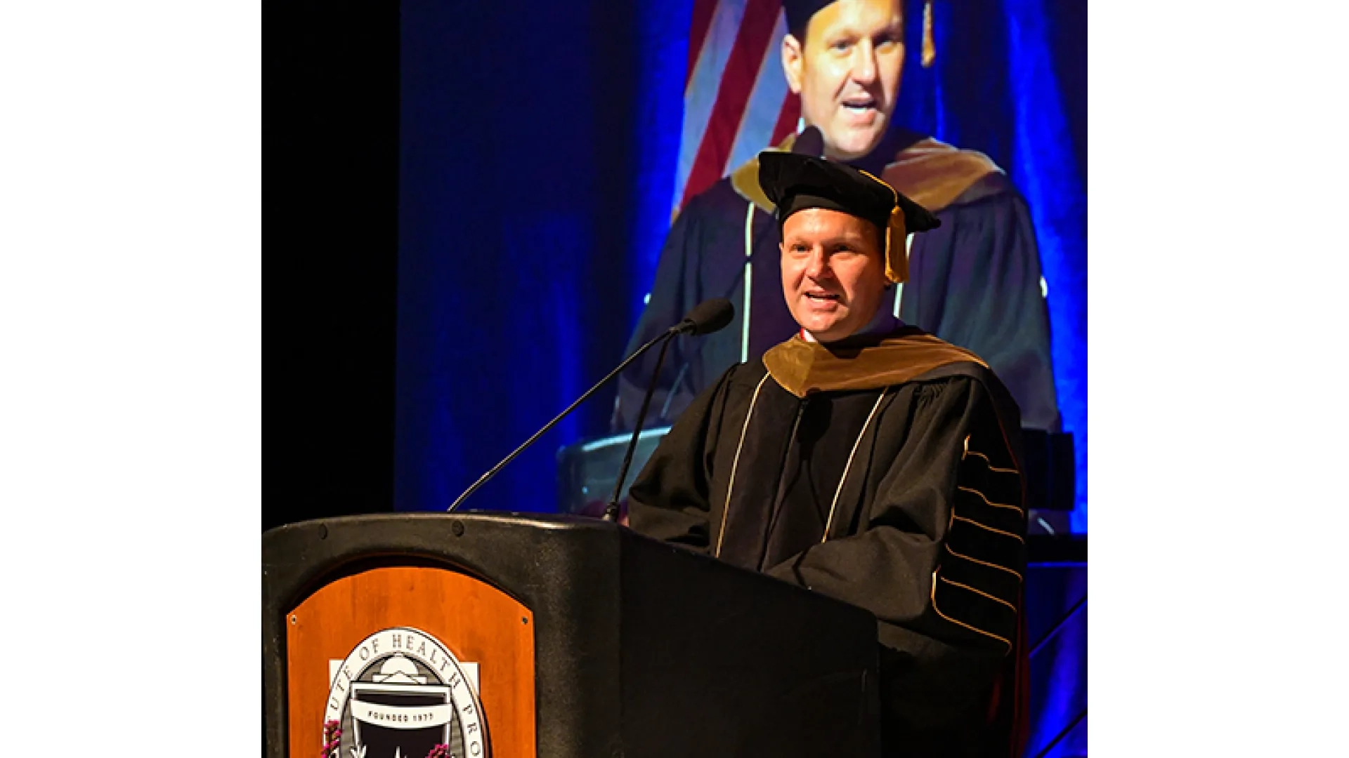 A man wearing a graduation gown and hat speaks at a podium at commencement