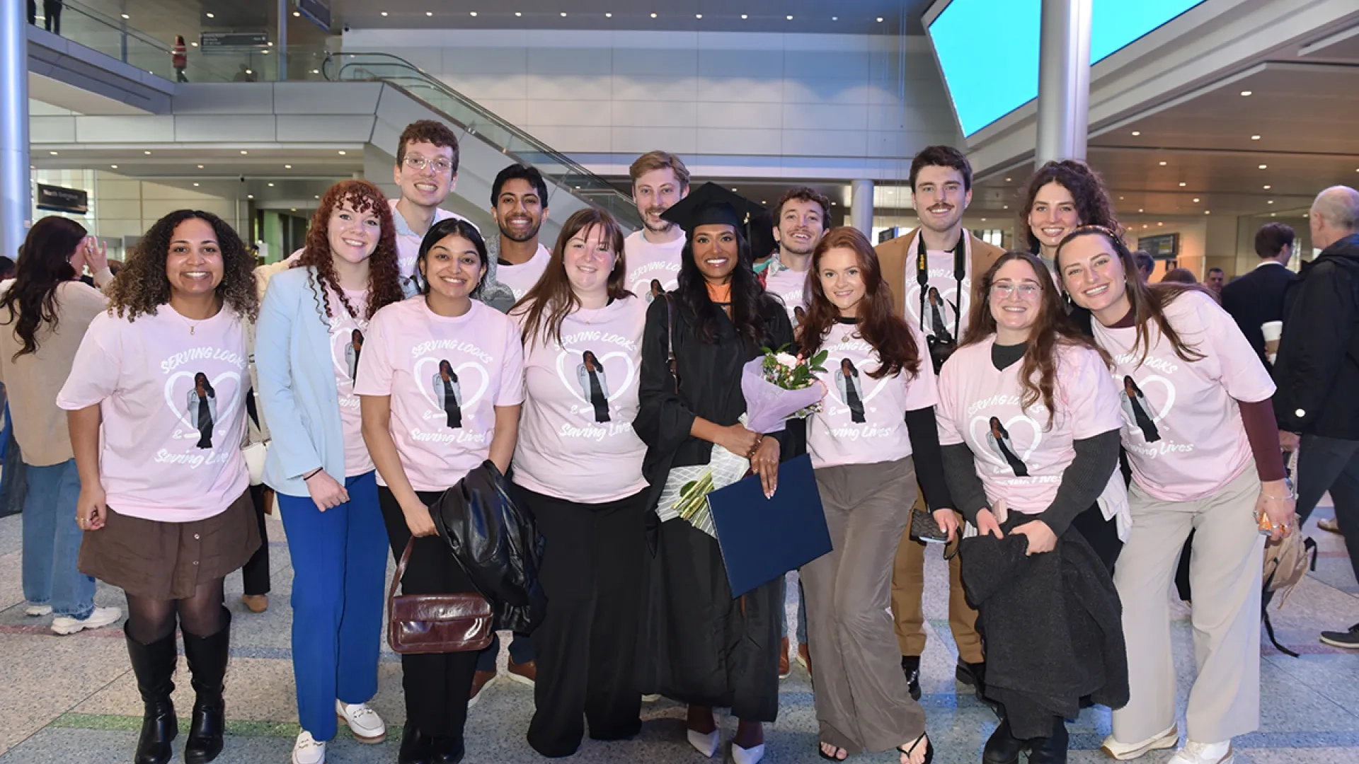 A group of people, one wearing graduation attire and several others wearing matching T-shirts, pose for a photo. 