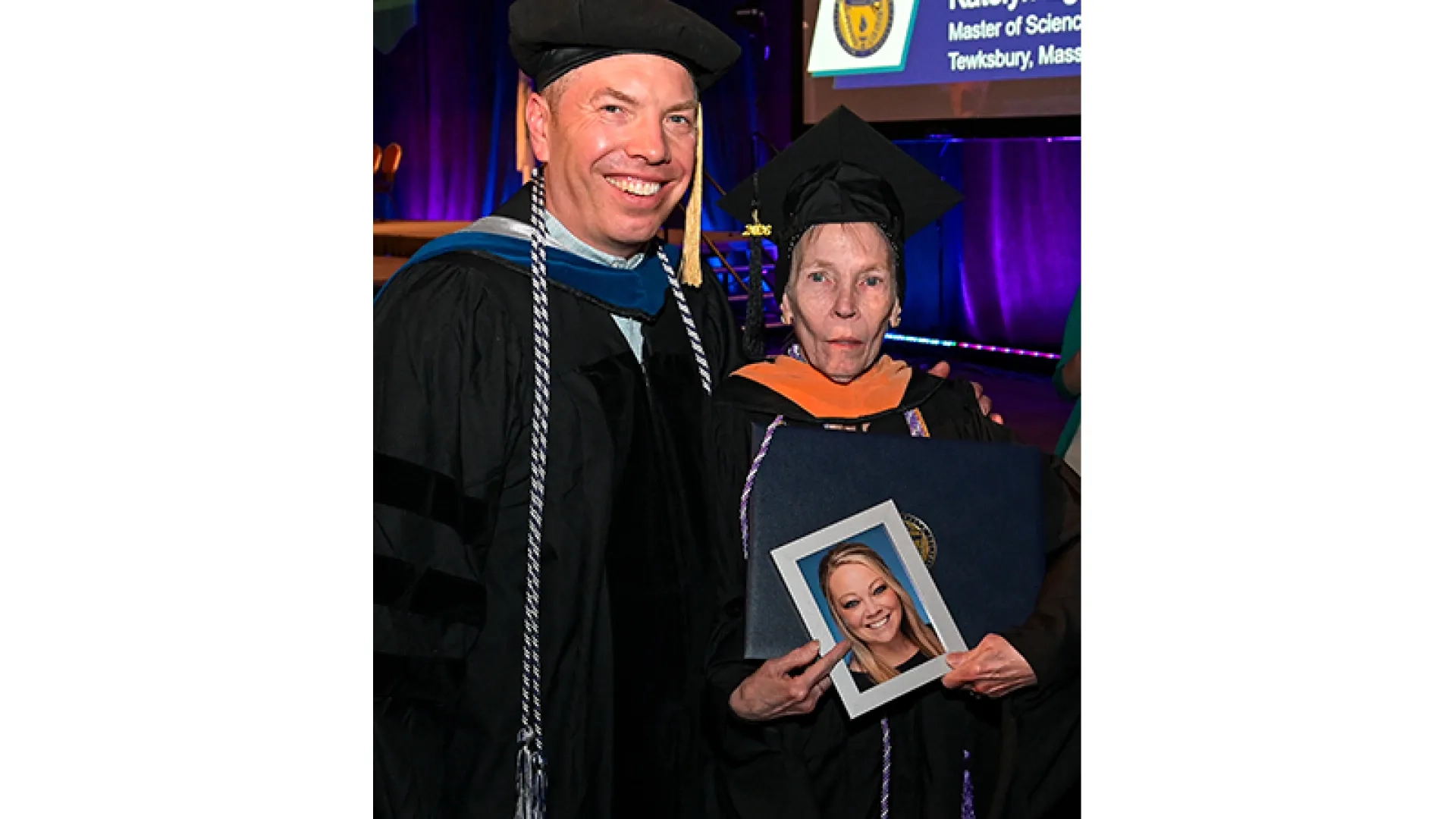 A man and woman in graduation attire stand next to each other with the woman holding a photo of a young woman