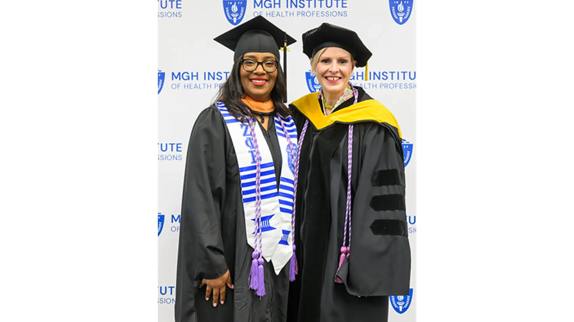 Two women in graduation gear pose in front of a backdrop that says MGH Institute of Health Professions