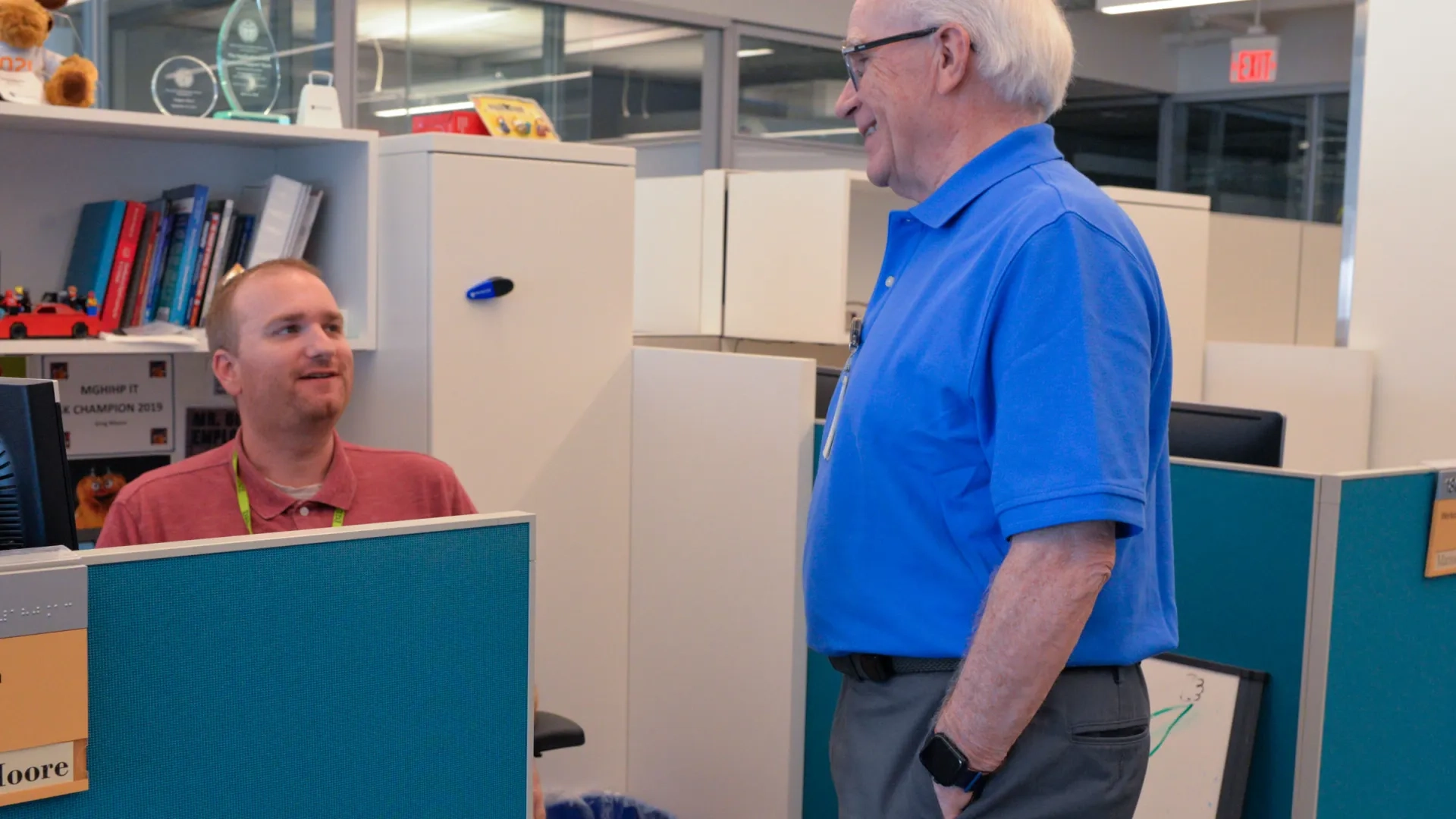 man sitting at desk talking to another man standing above the desk
