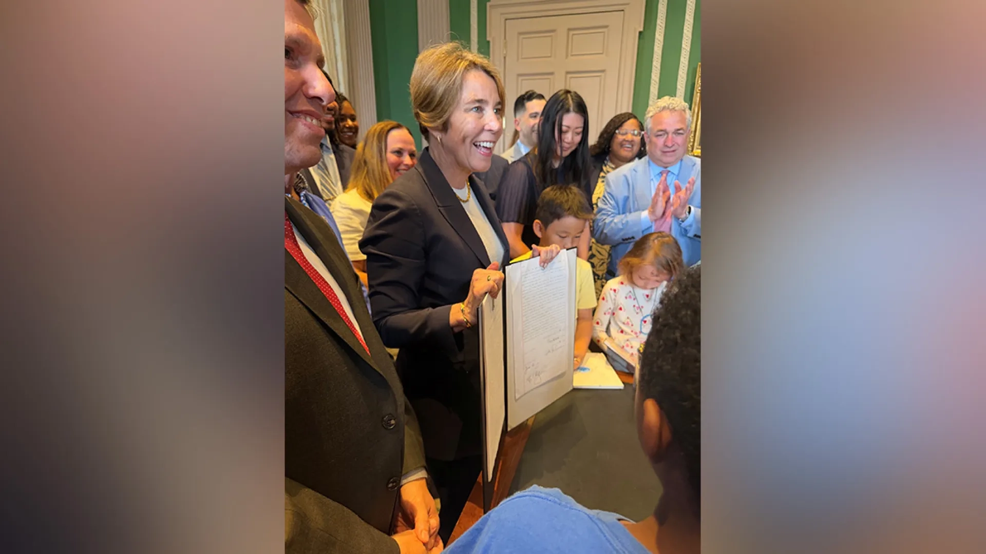 Governor Healey stands with a folder facing outwards with people gathered behind her