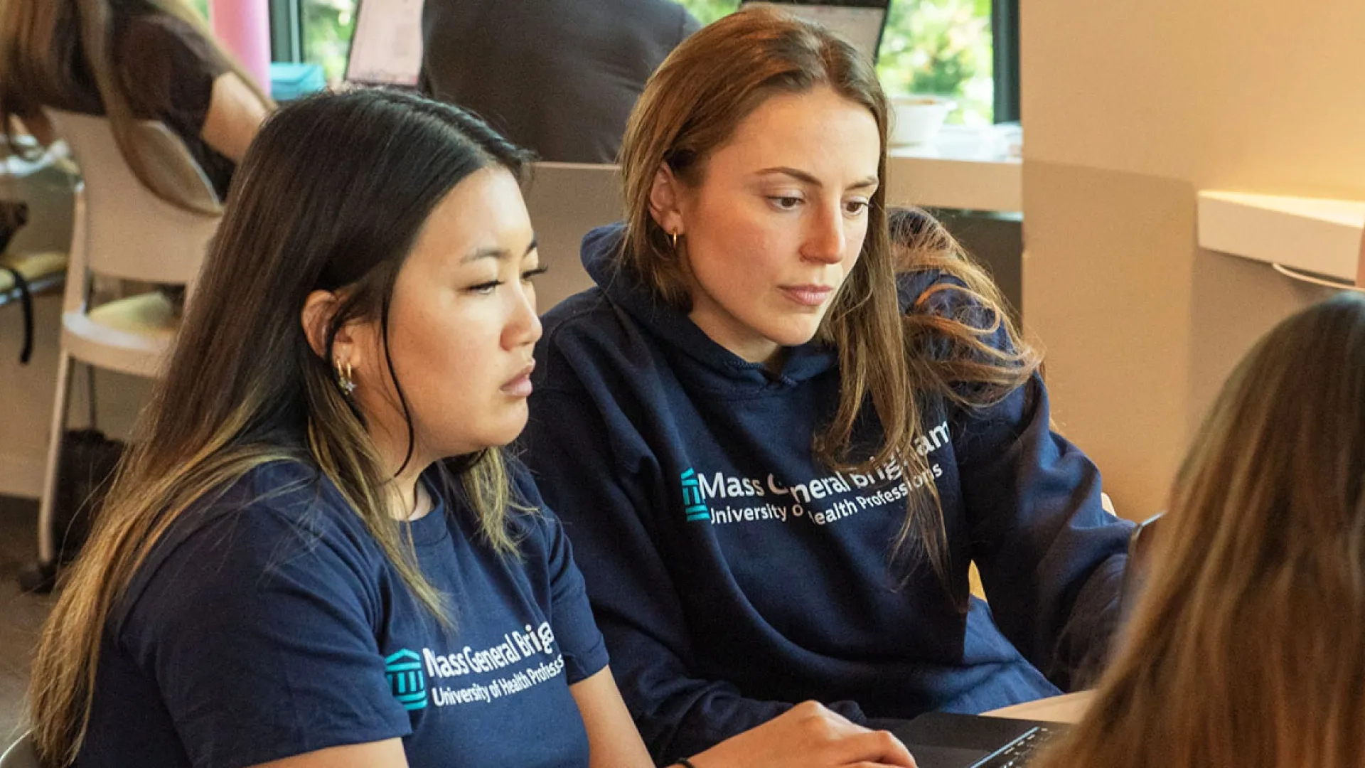 Two Mass General Brigham University students work during class wearing T-shirts with the university's name