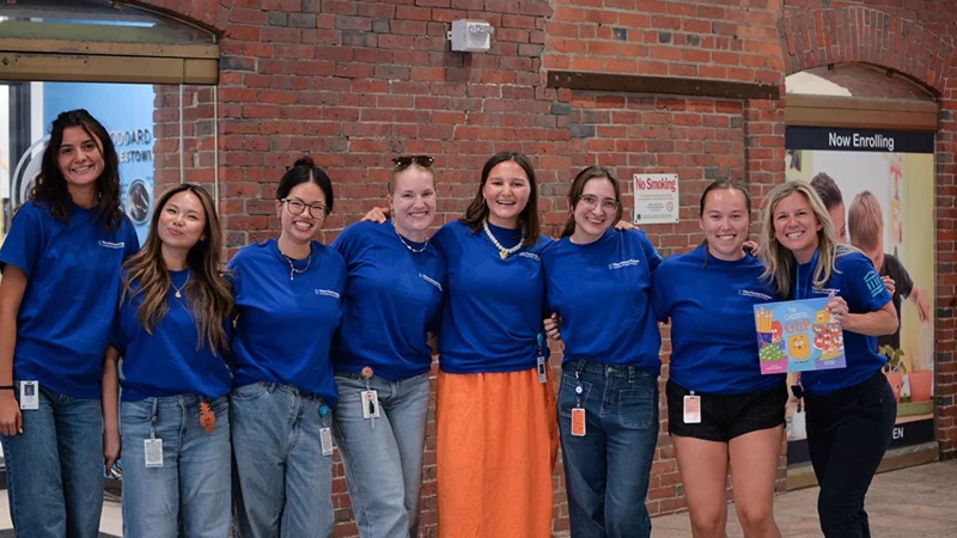 Volunteers in blue shirts posing for a photo in front of the Goddard School. One person is holding up a picture book.