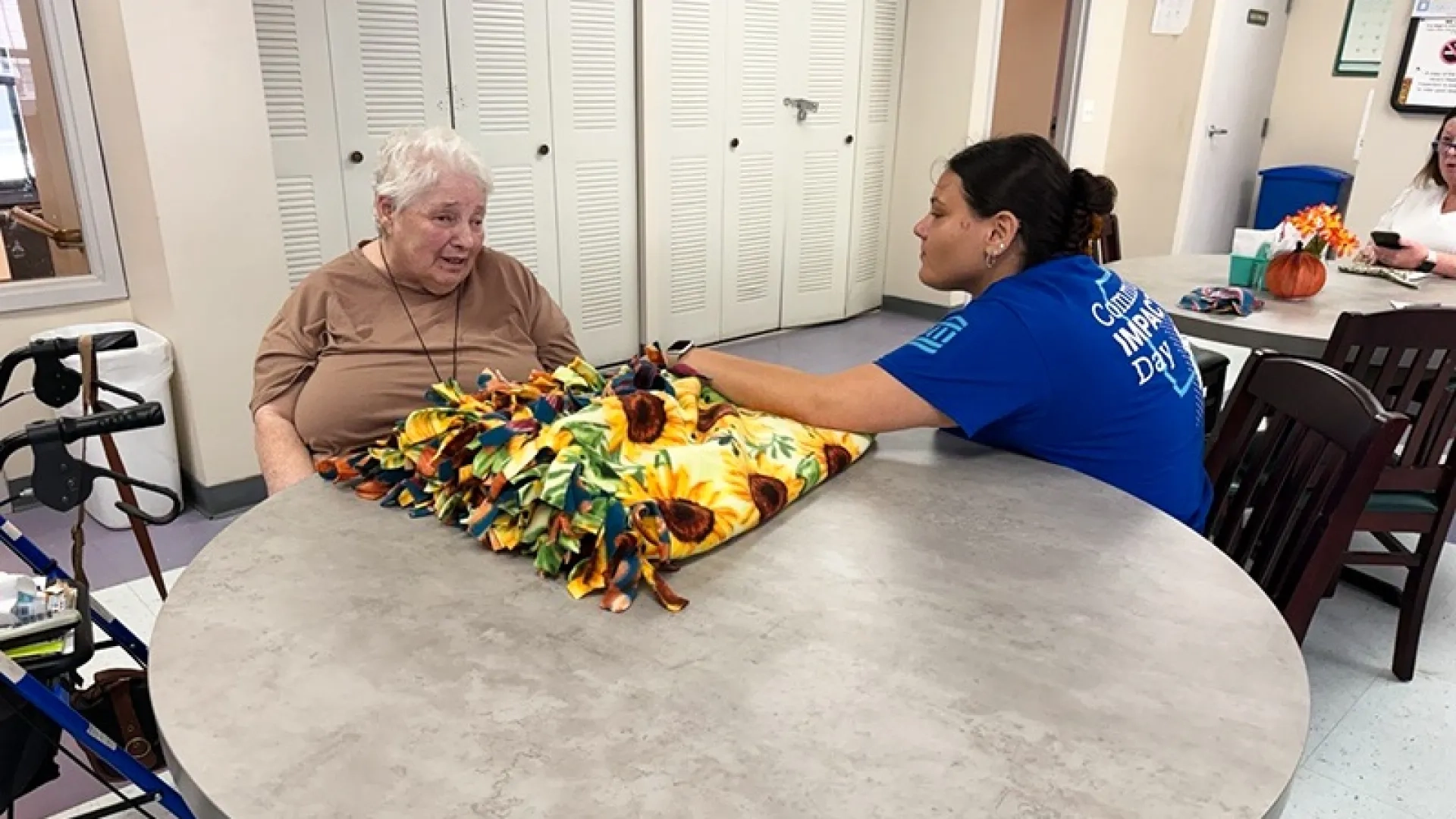 A woman in a blue shirt that reads "Community Impact Day" talks to an older woman in a nursing home. The older woman has a blanket with a sunflower pattern in front of her.