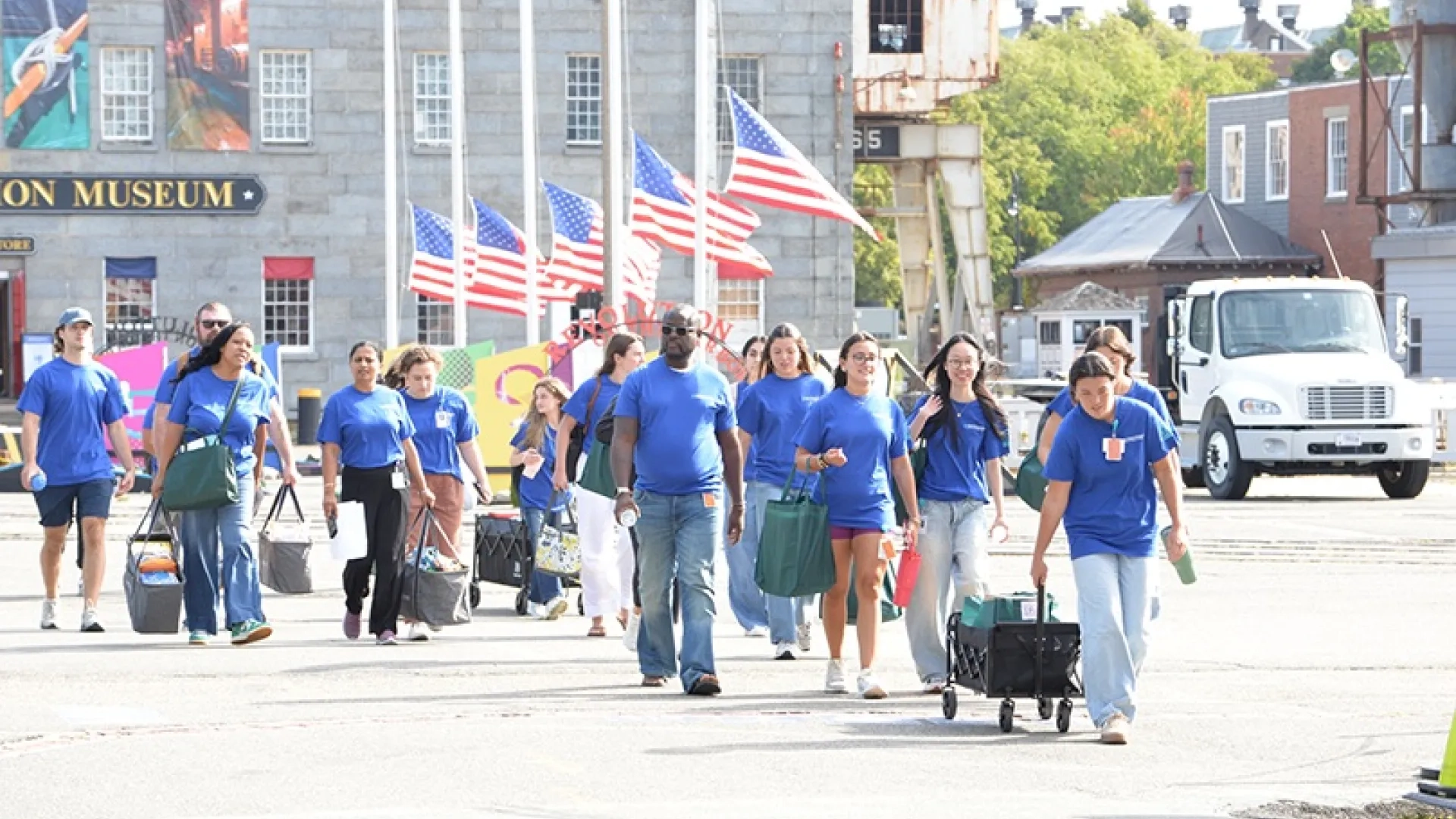Volunteers in blue t-shirts carry and wheel supplies in front of a building with American flags at half-mast