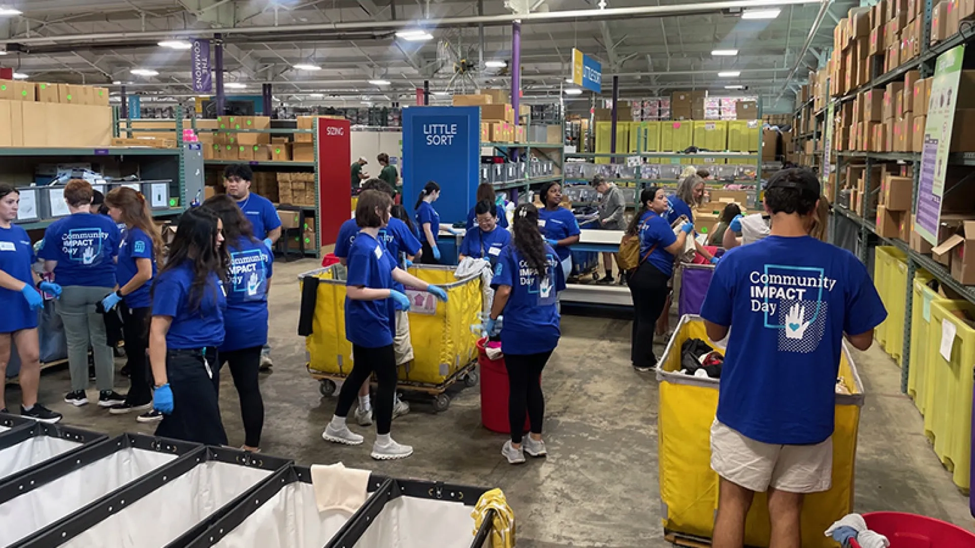 Volunteers in blue shirts that read "Community Impact Day" sort clothes and supplies into large yellow bins in a warehouse