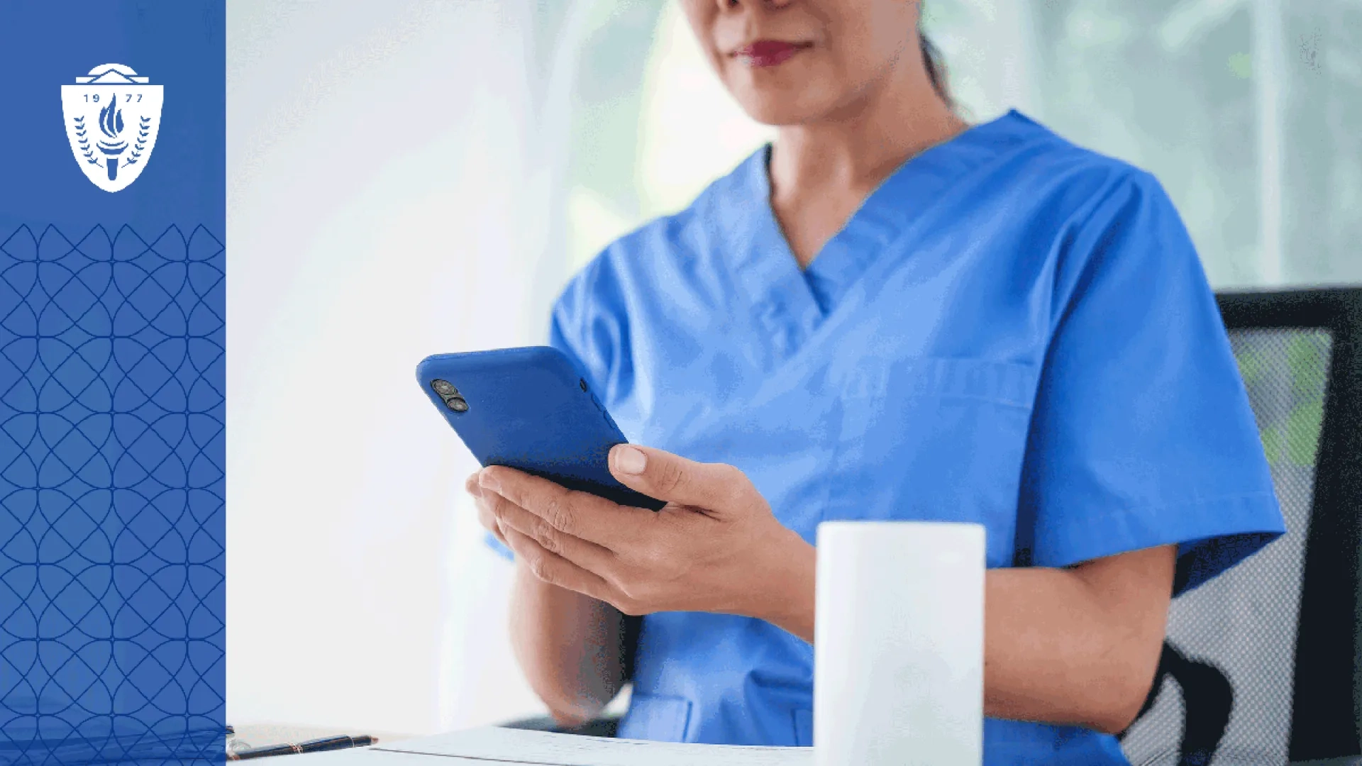 Woman in blue scrubs sitting a desk looking at her phone