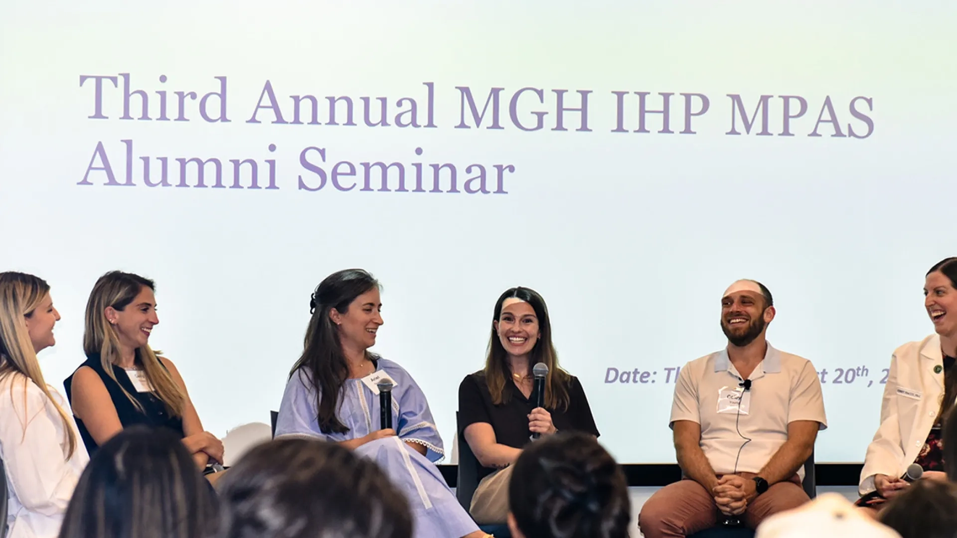A panel of alumni sits on a stage holding microphones with the words Third Annual MGH IHP MPAS Alumni Seminar beind them