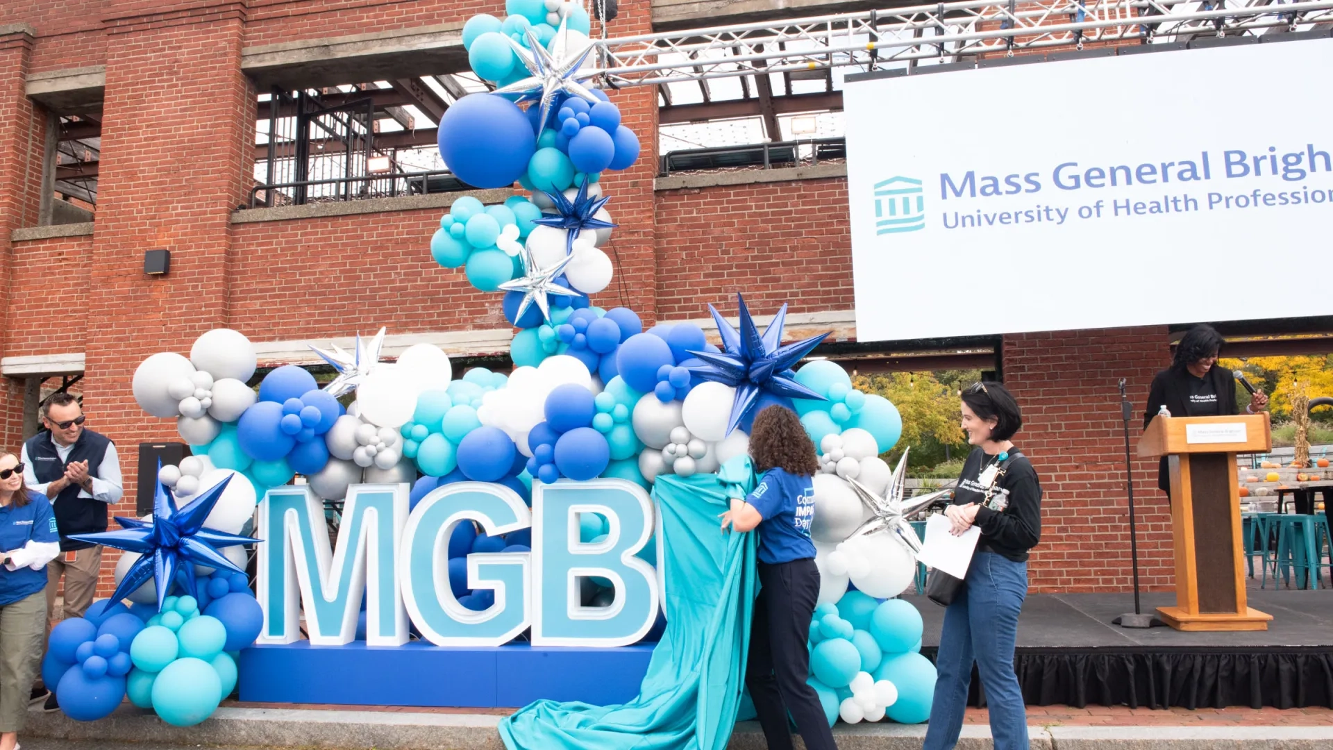Two women pulling a blue sheet off of letters that say MGBU