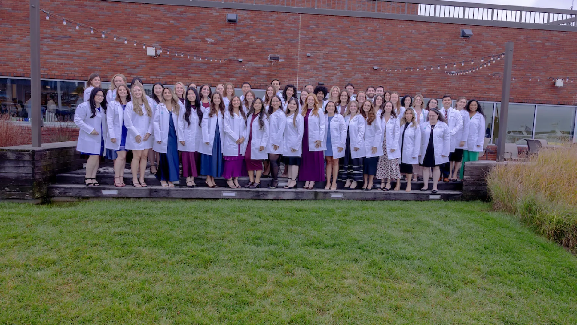 group photo of men and women in long white coats in front of a brick building with grass in front of them