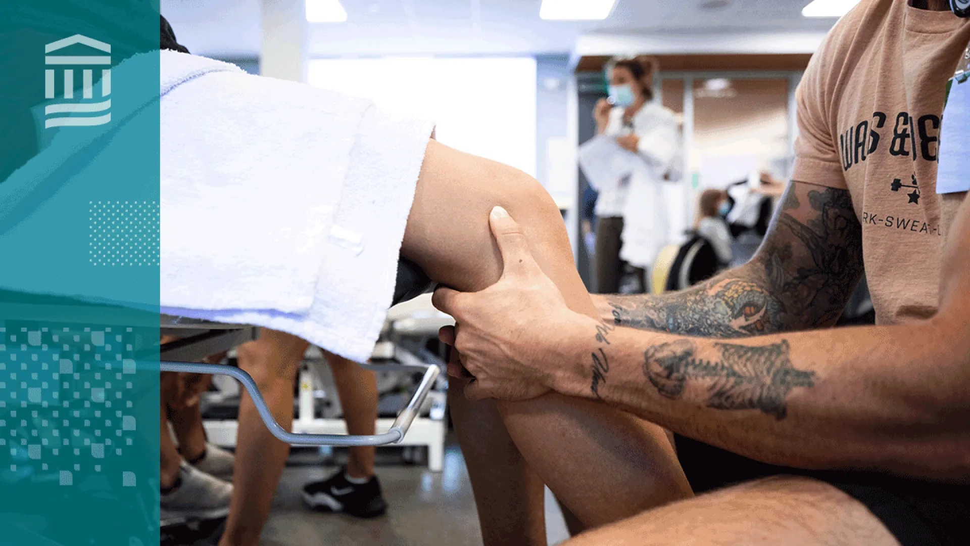a person sits in front of another who is on a medical table, the focus are the person's hands around the other's knee