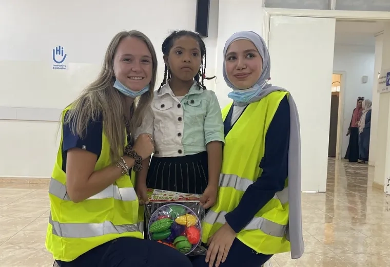 student in wearing yellow vest poses with a child and woman in a hijab also wearing yellow vest