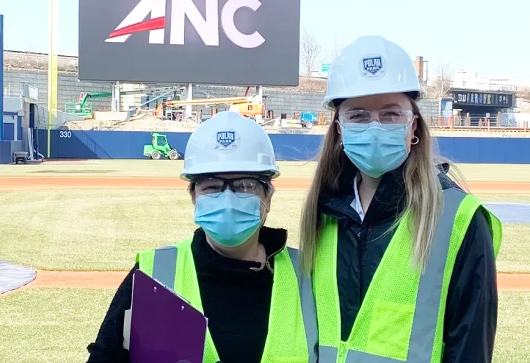 two women stand with ball field behind them wearing hard hats