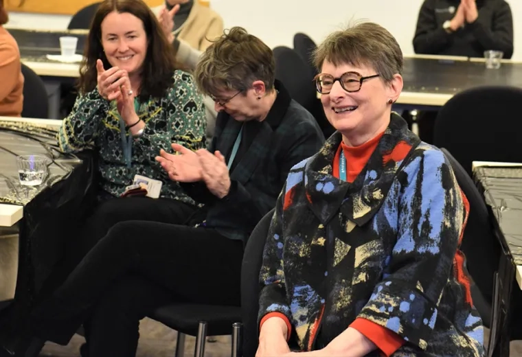 woman with glasses seated at a table smiles broadly as people clap around her