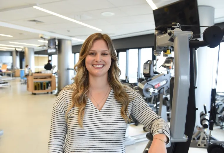 Tarryn has wavy long dark blonde hair and wears a black and white striped top as she leans against a weight machine in a gym