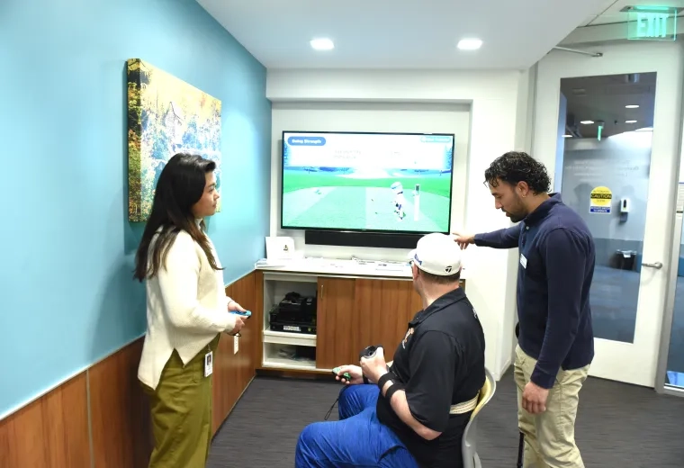 A man sits in a chair with a gaming controller in his hand looking at a television screen with a golf video game while another man points at the screen and a woman looks on
