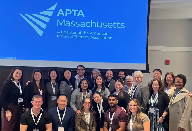 A group of people pose in front of a screen with the words APTA Massachusetts
