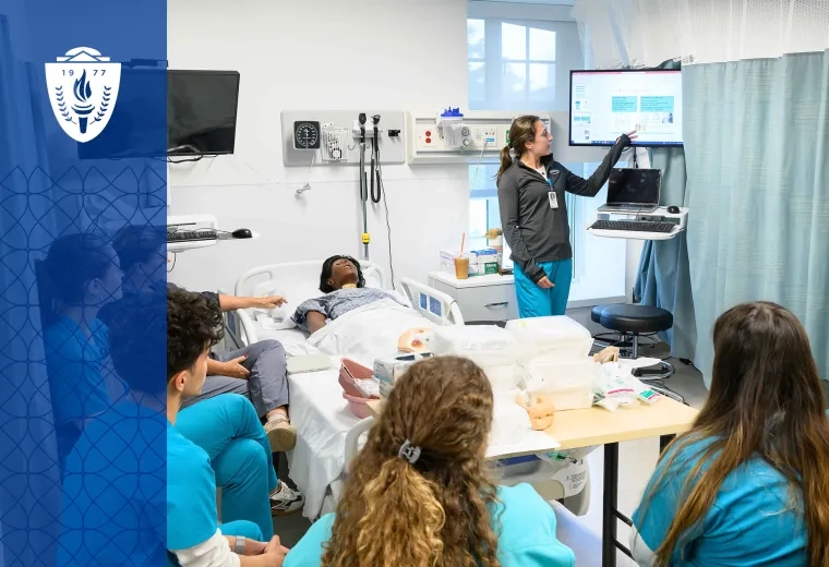 students in scrubs sit around a hospital bed containing a manikin while a woman points at a monitor above the bed