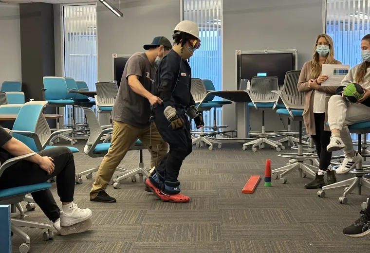 A person wearing a hard hat, goggles and gloves is assisted by another person while people watch