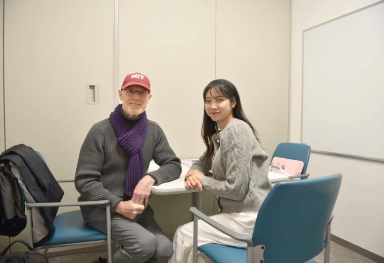 A man in a red baseball cap sits at a table facing the camera while a woman sits next to him facing the camera