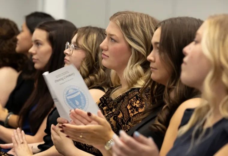 People sit in a row watching while one holds a piece of paper that says Pinning Ceremony