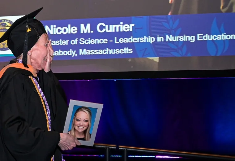 A woman walks across a commencement stage holding a photo of a young woman with a screen in the background with the name Nicole M. Currier